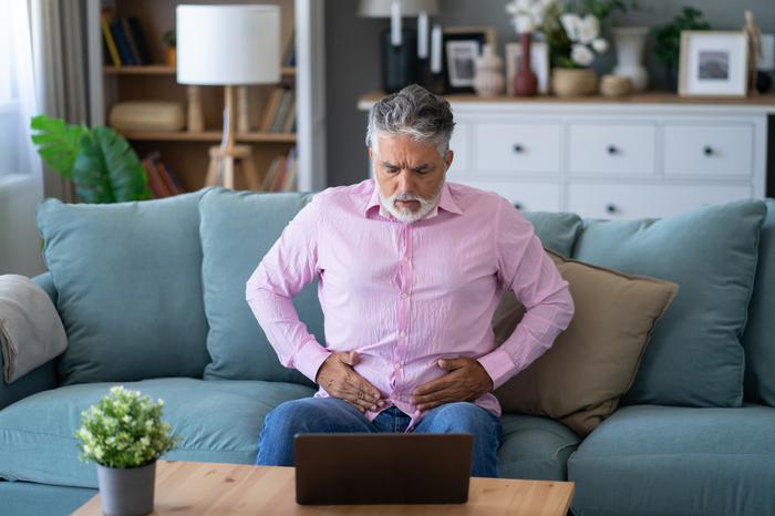 Patient sat on sofa at home and holding stomach whilst looking at laptop screen