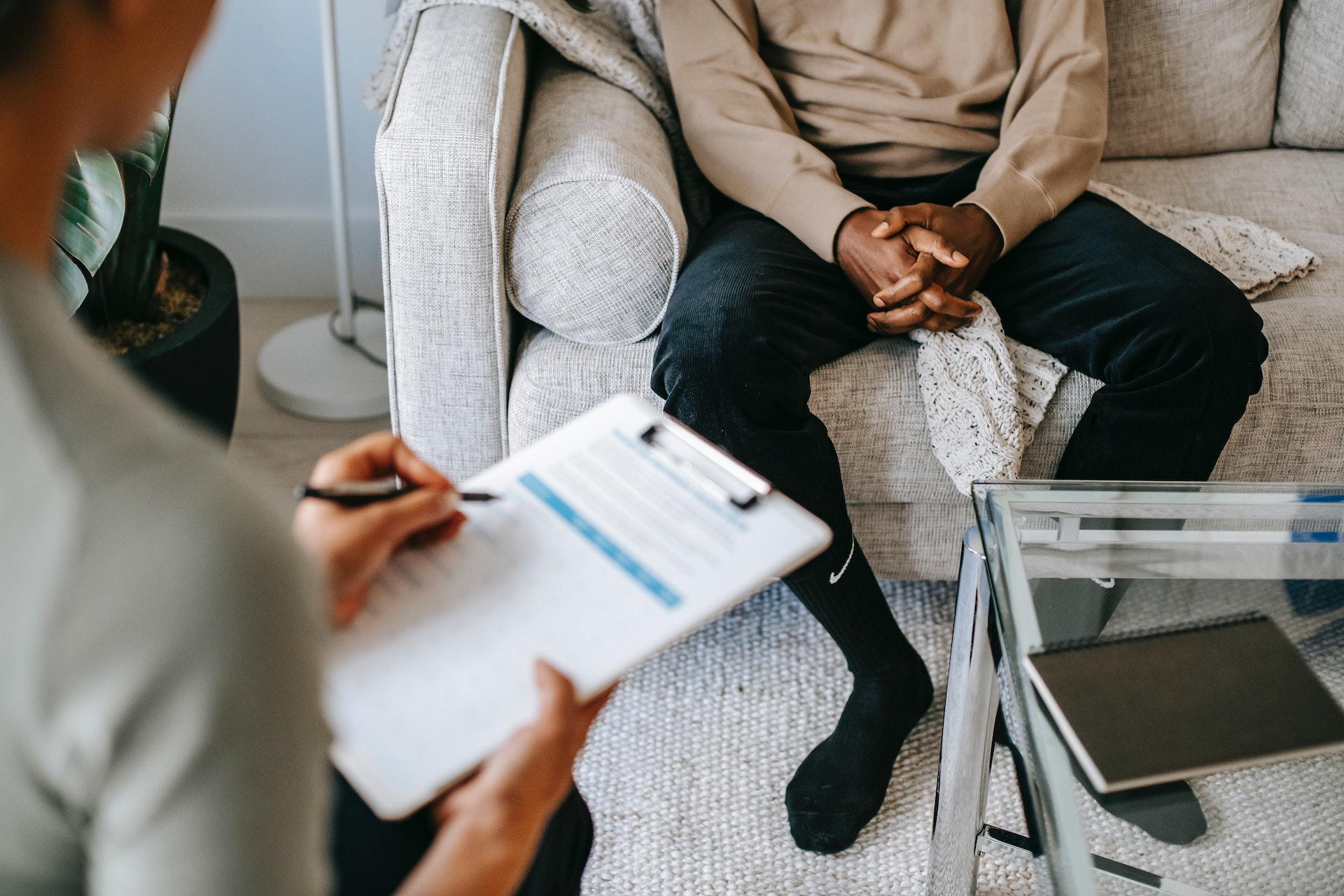 Black male patient sitting on a sofa and talking to a clinician, who is taking notes on a clipboard