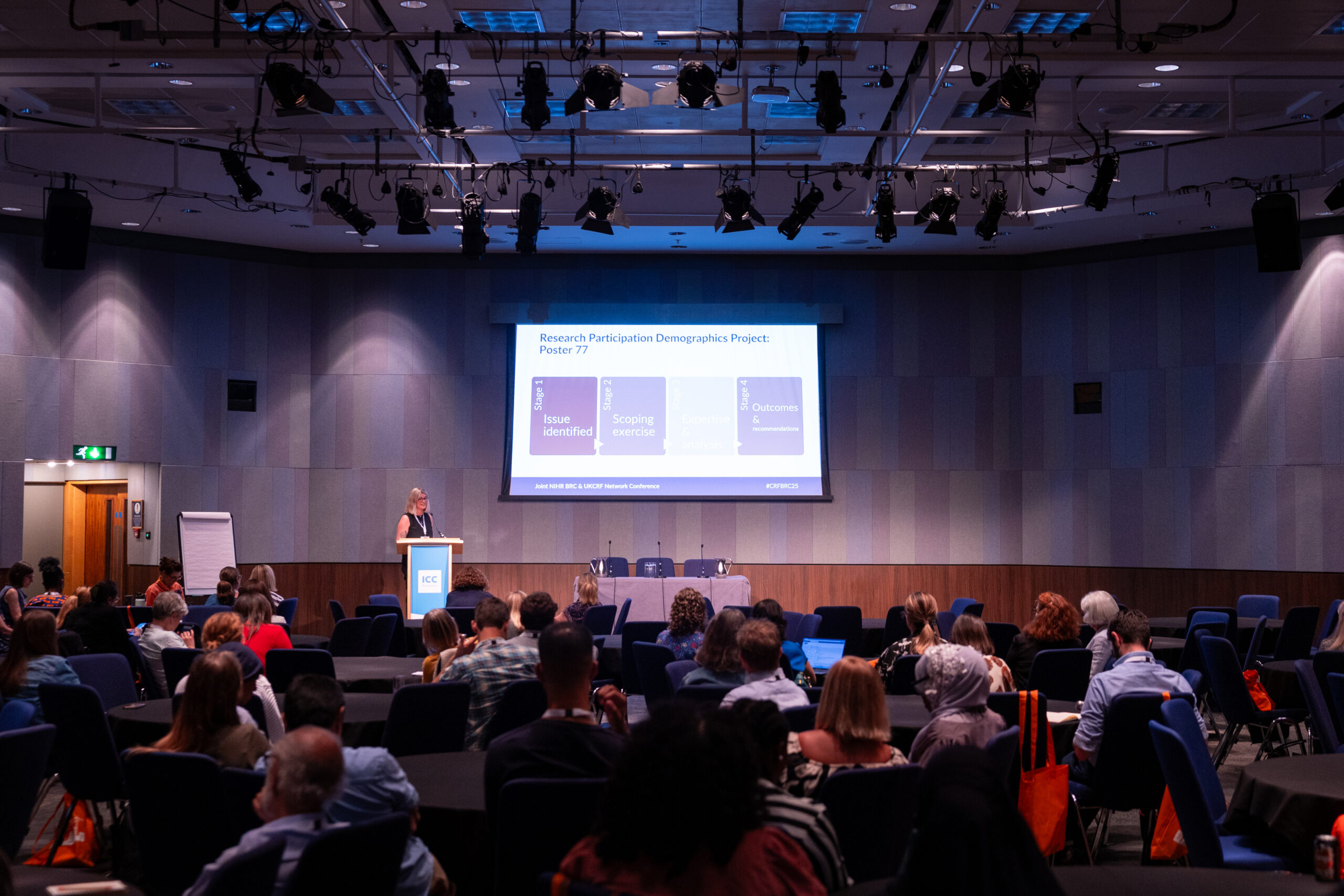 Medium size conference room with attendees sitting at tables and presentation being delivered at the front