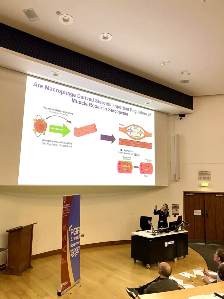 Woman presenting at a science symposium. On screen, her slide is titled 'Are macrophage derived steroids important regulators of muscle repair in sarcopenia'